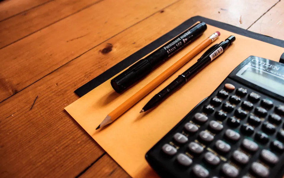 Calculator, pens and pencils resting on a paper envelope on a wooden desk