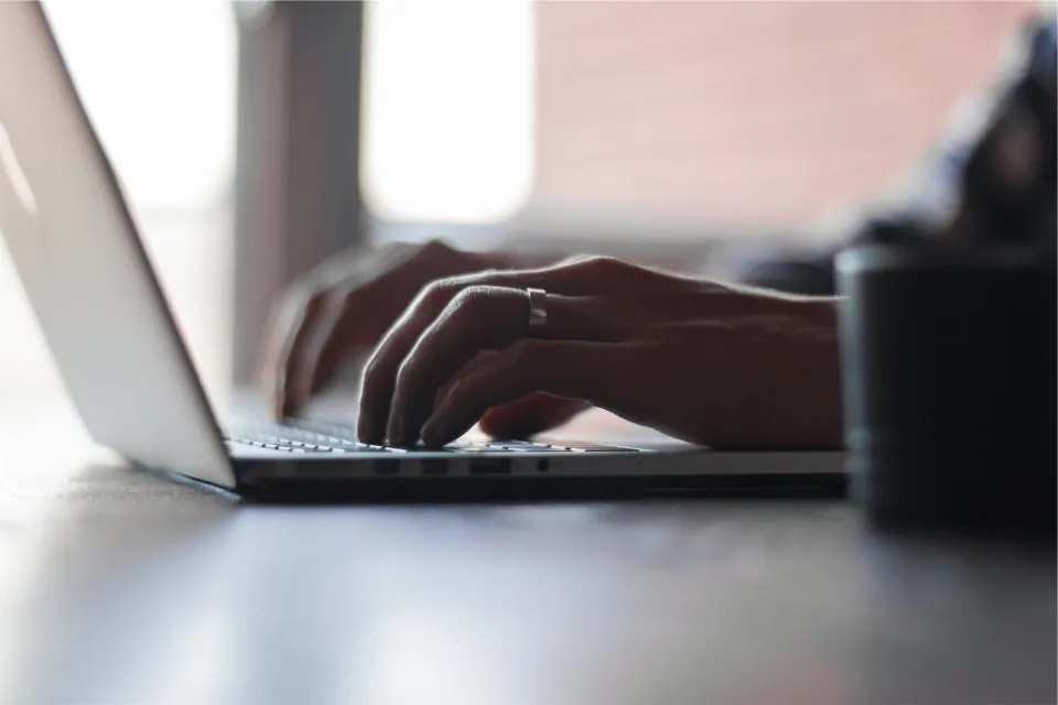 Hands typing on a laptop at a desk in soft window light