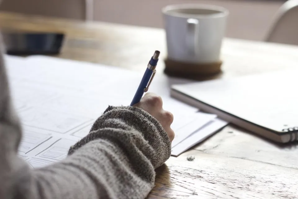 Person writing notes with a pen beside a coffee mug