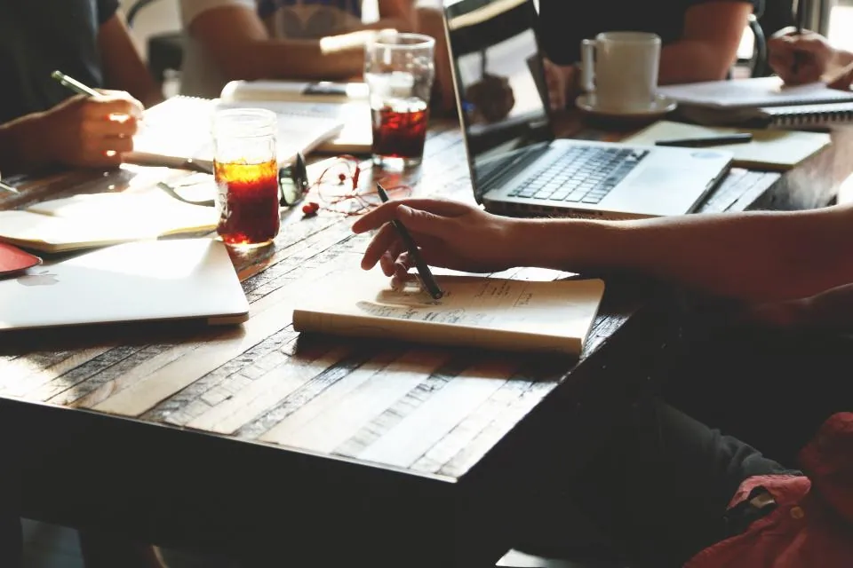 Coworkers around a wooden table with laptops, notebooks and drinks