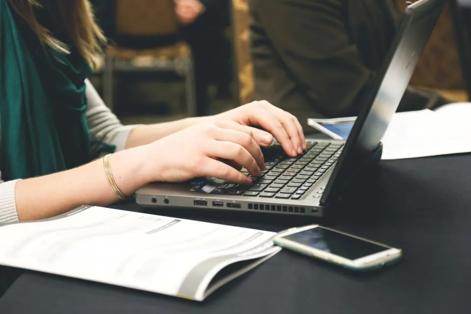Hands typing on a laptop beside a phone and an open notebook
