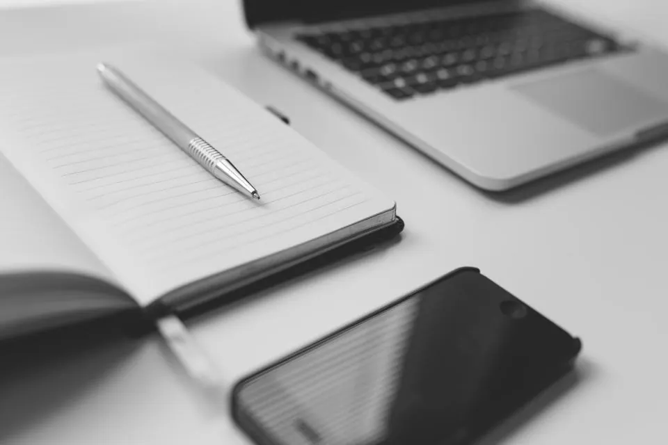 Open notepad with a pen next to a phone and a laptop