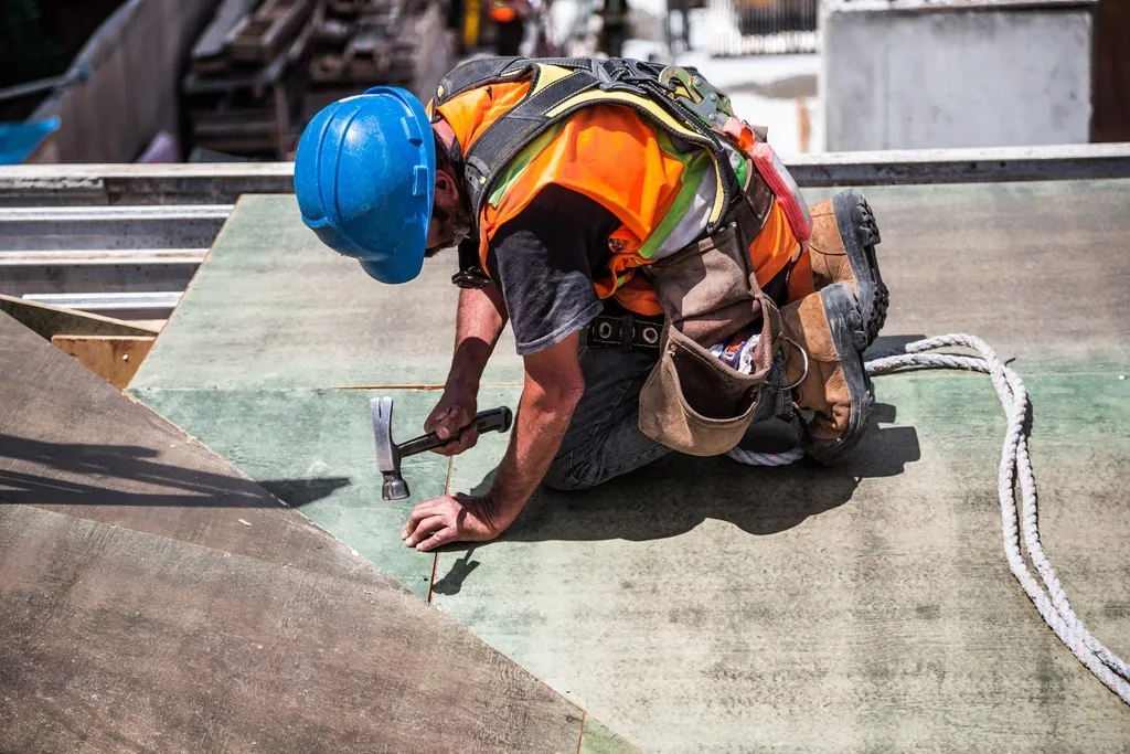 Roofer in a hard hat and safety harness hammering on a roof deck