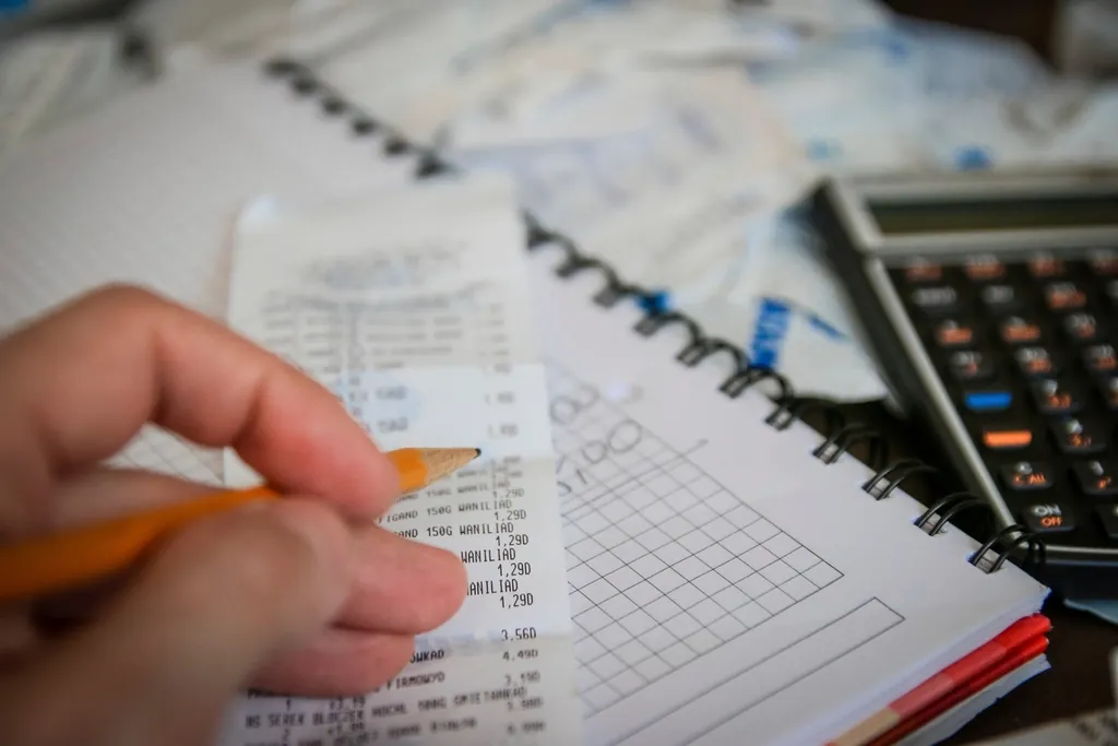 Hand with a pencil checking a receipt beside a notebook and calculator